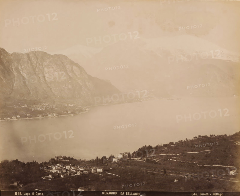 View of Menaggio from Bellagio, album page, photograph, This photo is part of an album., inscription: 'B28. Lado di Como, Menaggio da Bellagio, Ediz. Bosetti - Bellagio', annotation: '1898', photographer: Bosetti (possibly), Gotthard Pass, Bellagio (Italy), 1898, Lombardy, paper, albumen print, height 265 mm, width 350 mm, city-view in general, 'veduta', lake, mountains, Lake Como, Como, A sepia-toned panoramic view showing a large calm lake bordered by steep rocky mountains on one side and a gently sloping shoreline with scattered buildings and clusters of trees on the other, with a small village of tightly grouped houses in the foreground, terraced hillsides and winding roads, distant hazy peaks rising behind the nearer mountains, and a broad expanse of water occupying the central area of the image.