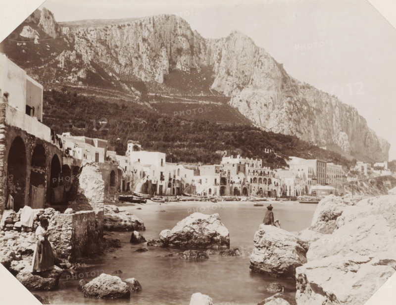 Island Capri, Capri la Marina Grande, View of the Marina Grande of Capri, photograph, Part of Album with 191 photos of a trip to and through South Africa., negative number: '2004', Capri, Isola di, publisher: Edizione Esposito, c. 1880 - c. 1920, photographer, paper, albumen print, height 198 mm x width 259 mm, harbour, A sepia-toned coastal scene showing a rocky shoreline and calm water in the foreground with large irregular rocks scattered along the shore and a person sitting on one rock facing the water. A woman stands on a rock near the left foreground wearing a long garment. Along the middle ground there is a row of arched stone structures and whitewashed buildings with balconies and doorways clustered at the water’s edge, and several small boats pulled up on the shore. Behind the buildings a terraced hillside rises with vegetation, and steep pale cliffs dominate the background, forming a high rocky mountain face that spans the image. The sky above is light and largely empty.
