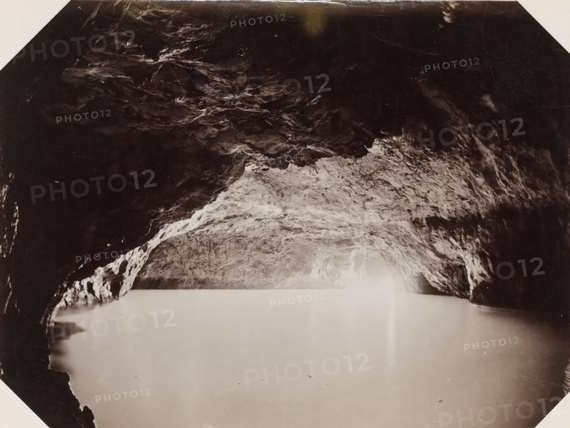 Capri la grotta azzurra preso dal vero, Interior of the Blue Grotto on Capri, Blue Grotto Capri, photograph, Part of Album with 191 photos of a trip to and through South Africa., negative number: '2094', Capri, Isola di, publisher: Edizione Esposito, c. 1880 - c. 1920, photographer, paper, albumen print, height 198 mm x width 258 mm, cave, grotto, A dark, irregular rocky ceiling with textured surfaces fills the upper portion of the image, transitioning to a lighter, rough rock surface toward the center where uneven striations and small protrusions are visible; the lower area shows a smooth, pale expanse with subtle reflections and faint speckles, and darker rock formations frame the left and right edges creating an archlike opening above the lighter central area.