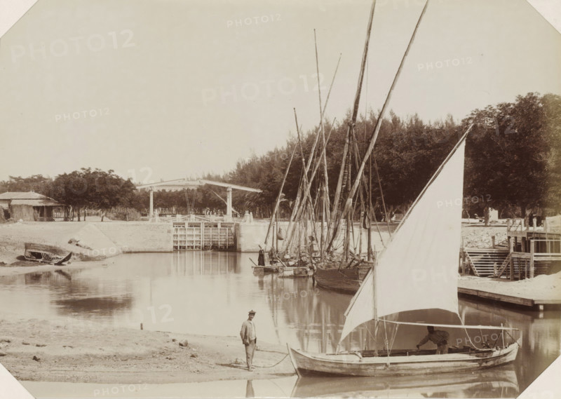 Sailing ships at a Suez Canal lock, Suez Canal, photograph, Part of Album with 191 photos of a journey to and through South Africa., c. 1870 - c. 1890, photographer, North Africa, paper, albumen print, height 195 mm x width 280 mm, canal, sluice, weir (landscape with figures, staffage), A calm river scene showing several sailboats moored along a bank, their tall masts clustered together and reflected in the water, a large triangular sail in the foreground partially obscuring a boat where a person leans over the gunwale, a second person standing on the sandy shore holding a rope, a small beached boat resting on the far bank, a wooden lock or gate structure spanning the water with a simple overhead mechanism, a cluster of trees and rustic buildings behind the bank, and wooden steps and a small pier area adjacent to the boats.