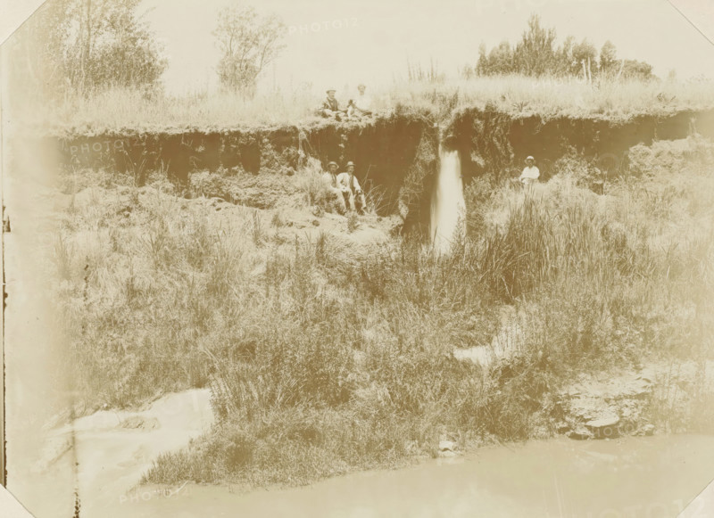 Near Pretoria, Europeans with a dog next to a waterfall near Pretoria, photograph, Part of Album with 191 photographs of a trip to and through South Africa., c. 1890 - c. 1920, photographer, paper, daylight gelatin silver print, height 213 mm x width 293 mm, waterfall, Pretoria, A pale sepia photograph showing a grassy riverbank with a vertical eroded cliff face and a narrow waterfall flowing from the top to the lower vegetation, tall grasses and reeds growing along the bank, scattered bushes and small trees behind the cliff, and five seated people wearing hats and light clothing positioned along the top and middle of the bank, with one person sitting alone near the right edge and others grouped nearer the center, all surrounded by natural terrain and vegetation.