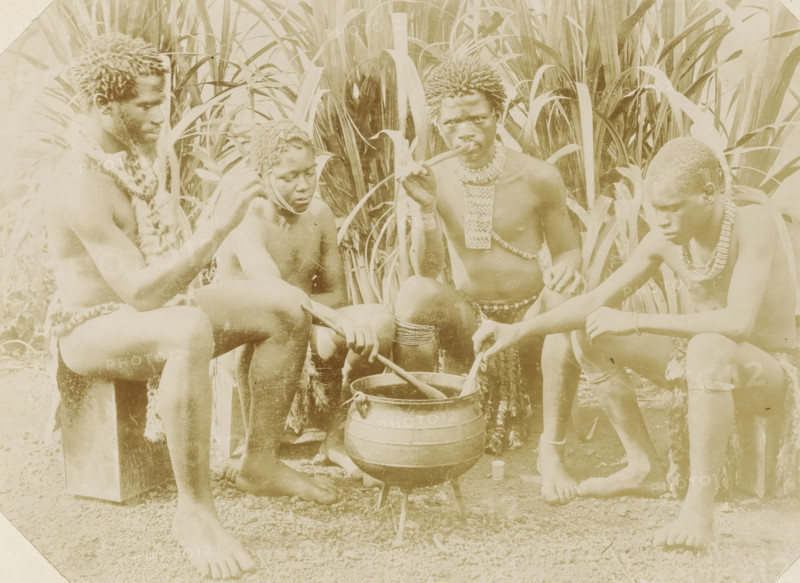 Four black men sitting around a cooking pot in South Africa, Kaffirs, photograph, Part of Album with 191 photographs of a trip to and through South Africa., South Africa, c. 1890 - c. 1920, photographer, paper, daylight gelatin silver print, height 213 mm x width 292 mm, Africans, kitchen-utensils: pot, Four shirtless people sit close together in front of tall, broad-leafed plants surrounding them. They all have short, textured hair and wear decorative necklaces and waist adornments made from beads and natural materials. One person on the left wears a fur-like garment draped over a shoulder and sits on a block while looking toward the center. Next to them, a person with beaded strands near their face looks downward while holding a stirring stick. In the center, a person holds a long pipe to their mouth and has layered necklaces and bracelets, with one hand resting on a knee. On the right, a person leans forward concentrating on stirring a large pot set on a small tripod over the ground...