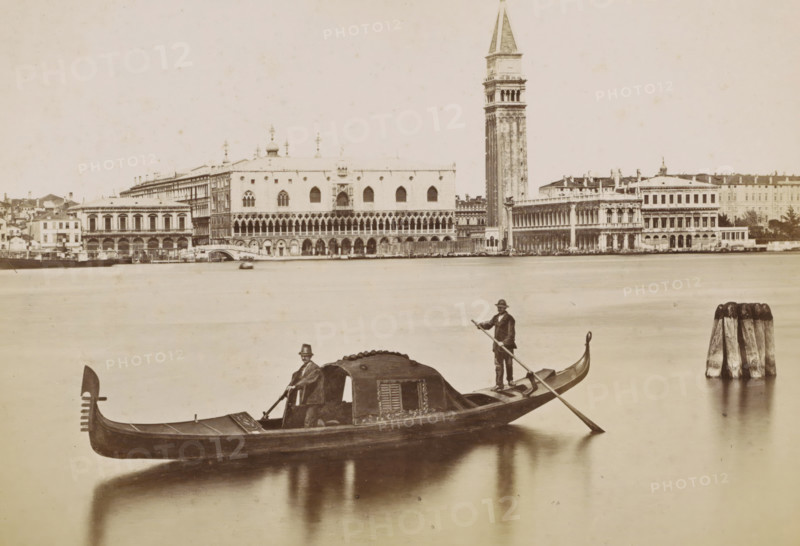 View of the Doge's Palace, the Campanile and surrounding buildings in Venice with a gondola in the foreground, Venezia - panorama e gondola, photograph, Part of Topographical album of Venice 1881., check stamp: 'Carlo Ponti Venezia', negative number: '1', photographer: Carlo Ponti, Venice, 1860 - 1881, Carlo Ponti, photographer, paper, albumen print, height 199 mm x width 258 mm, prospect of city, town panorama, silhouette of city, canals, waters (in city), palace, tower, clock tower, monumental adornment, gondola, Roman Catholic Church, travel, Doge's Palace, St. Mark's Square, Campanile di San Marco, A sepia-toned waterfront scene showing a gondola in the foreground with a covered cabin and two men, one seated near the cabin and one standing at the stern holding a long pole, their reflections visible in the calm water; the background features an ornate riverside building with a colonnaded facade and arched windows, a tall bell tower rising beside it, rows of adjacent classical buildi...