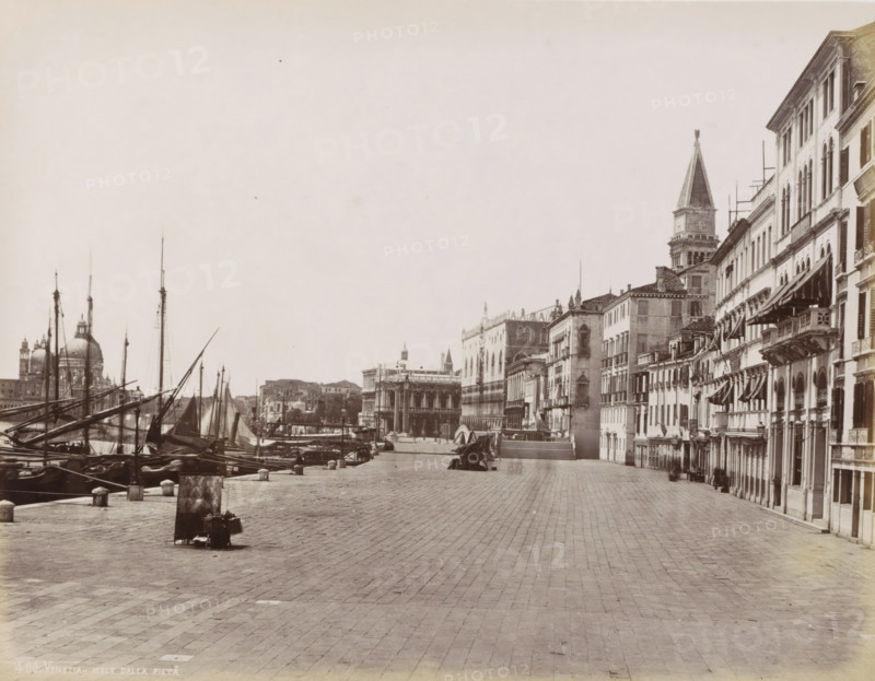 Quay in Venice seen towards St. Mark's Square, Quai des Esclavons à Venise, Venezia - Molo dalla Pietà, photograph, Part of Topographical album of Venice 1881., negative number: '400', check stamp: 'Carlo Ponti Venezia', photographer: Carlo Ponti, Venice, 1860 - 1881, Carlo Ponti, photographer, paper, albumen print, height 200 mm x width 259 mm, quay (city(-scape) with figures, staffage), tower, clock tower, monumental adornment, Roman Catholic Church, travel, A wide riverside promenade paved with large rectangular stones runs along the right side of the image, bordered by a row of multi-story buildings with arched windows, balconies, and striped awnings, while to the left several moored wooden boats with tall masts and rigging line the water's edge; in the distance a domed church and a tall bell tower rise above the cityscape, and a colonnaded building stands near the waterfront, all under a pale sky.