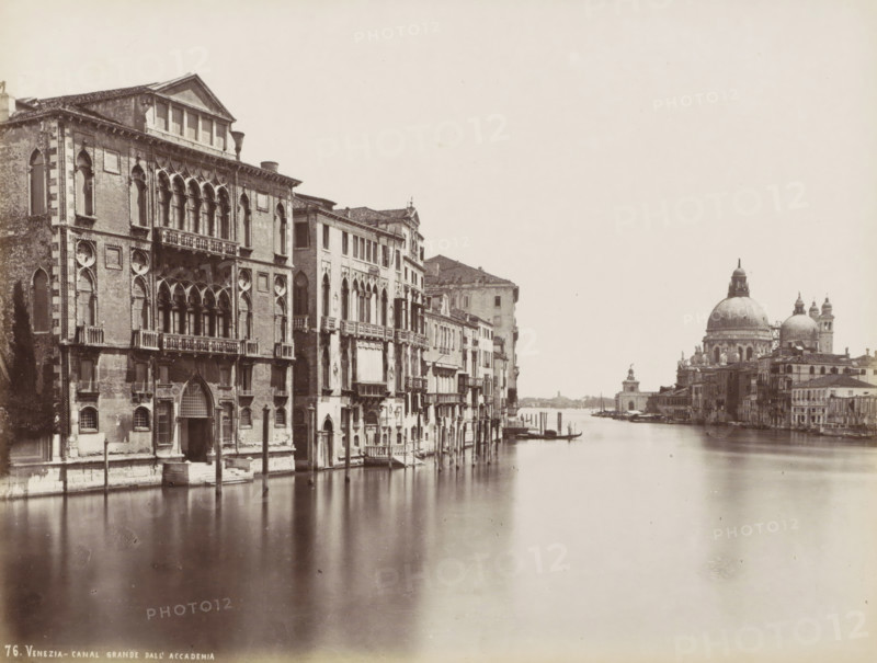 Venezia - Grand Canal from the Accademia, Grand Canal., Grand Canal with palaces, the Basilica of Santa Maria della Salute and the Fondamente Salute in Venice, photograph, Part of Topographical Album of Venice 1881., negative number: '76', check stamp: 'Carlo Ponti Venezia', photographer: Carlo Ponti, Venice, 1860 - 1881, paper, albumen print, height 200 mm x width 260 mm, canal, Roman Catholic Church, travel, Basilica of Santa Maria della Salute, Grand Canal, A riverside scene showing a wide waterway bordered by a row of ornate, multi-story brick and stone buildings with arched windows, balconies, decorative facades, and visible shutters on the left, wooden poles rising from the water along the building fronts, small boats moored at the edges, and a distant waterfront skyline on the right dominated by a large domed church with smaller domes and bell towers, all rendered in a monochrome photographic tone.