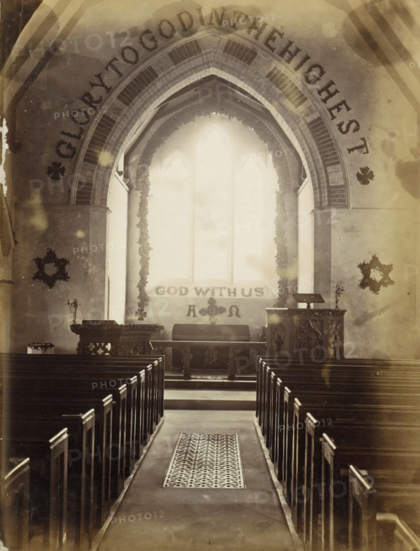 Xmas 1864, Interior of S. Peter's Church Sea View, Interior of St. Peter's Church in Seaview on the Isle of Wight during Christmas, photograph, Part of an English family album with photos of people, travel, cricket and artwork., Seaview, 1864-12-25, photographer, Isle of Wight, paper, albumen print, height 205 mm x width 162 mm, parts of church interior: nave, parts of church interior: choir, A central aisle with a patterned runner leads between rows of wooden pews toward an elevated chancel area framed by a large pointed arch with decorative brickwork, flanked by two tall vertical panels and garlands hanging down each side of a bright triple-lancet window, with an altar and ornately carved wooden pulpit on either side and candle holders visible, wreath-like decorations on the side walls, and a tiled step and low railing at the front.