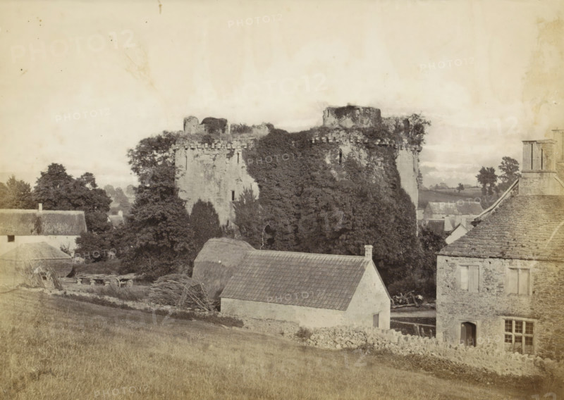 Nunney Castle Somerset, View of Nunney Castle and the surrounding buildings, photograph, Part of an English family album with photos of people, travel, cricket and artwork., Nunney, c. 1860 - c. 1870, photographer, Somerset, paper, albumen print, height 158 mm x width 208 mm, castle, ruin of a dwelling, house, plants and herbs: ivy, A sepia-toned scene showing a stone ruin partially covered in dense foliage rising behind a group of buildings, the ruin featuring battlemented tops and a round tower section. In front of the ruin, there are trees and hedges interspersed among a cluster of houses with pitched roofs, one roof tiled and another thatched, and a stone house with visible windows and a doorway. In the foreground a sloping grassy field leads down toward a low stone wall and scattered farm materials, including bundled branches and a wooden cart wheel. The sky above is pale and expansive, occupying the upper portion of the image.