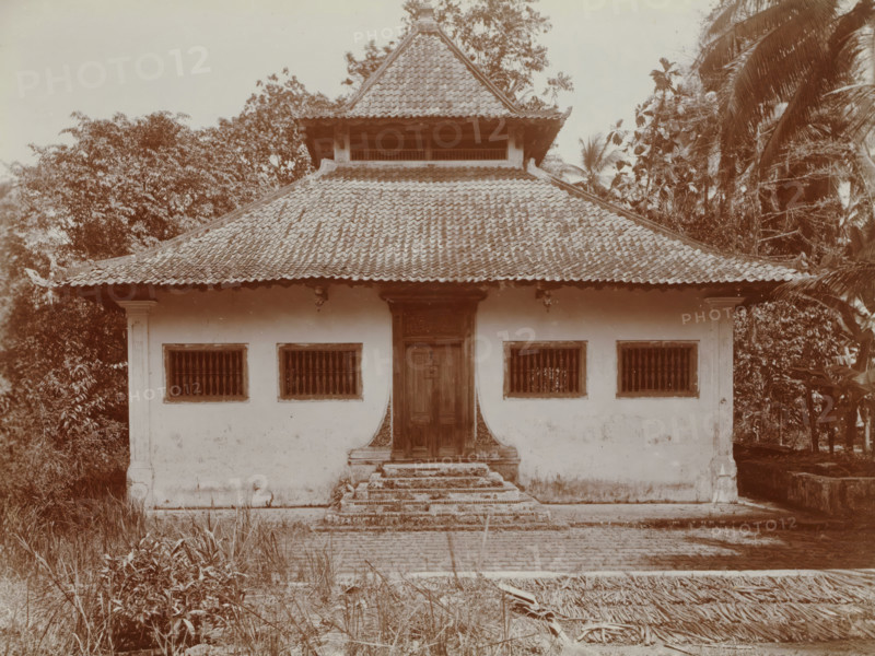 Missigit at Angké. No longer in use., Exterior of a mosque at Angké (Batavia), album page, photograph, Part of Travel Album with photos of Batavia., Batavia, c. 1920, photographer, Java, West Java, paper, daylight gelatin silver print, height 248 mm x width 305 mm, temple, shrine, Islam, Mohammedanism, overseas history, colonial history, Dutch East Indies, The, A single-story building with a tiled multi-tiered roof and a small upper pavilion sits centered in the image, flanked by dense trees and tall palm fronds on both sides; the building facade shows a central wooden door accessed by worn stone steps and framed by decorative trim, with two barred windows symmetrically placed on either side set into a smooth light-colored wall, and small exterior light fixtures under the roof eaves; the foreground contains overgrown grasses and scattered plant debris leading to a paved area in front of the steps.