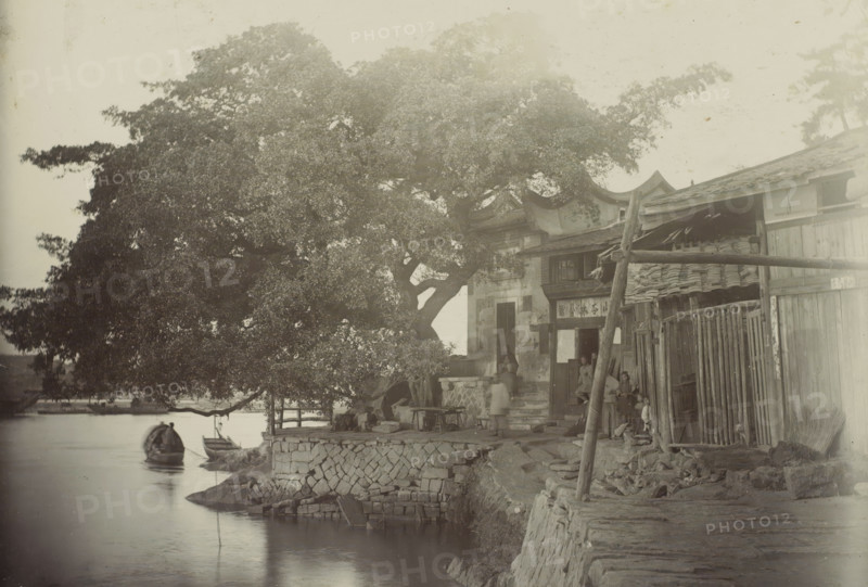 View of a house with a yard along the water, presumably in Fuzhou, photograph, Part of travel album by H. Dunlop with photographs of sights in Persia, Scotland, Germany, Russia, China, and Canada., photographer: Hendrik Dunlop (possibly), Fuzhou, c. 1890 - c. 1895, Fujian, paper, daylight gelatin silver print, height 106 mm x width 153 mm, courtyard, pier, quay, wharf, A riverside scene with a large leafy tree dominating the left and center, its branches extending over the water and a stone embankment. Small wooden boats float near the shore beneath the tree. Along the right side there are wooden buildings with tiled roofs and open doorways, a narrow stone path and steps leading up to one entrance. Several people are visible near the doorways and on the steps, some seated and some standing, with indistinct clothing and faces. Stacked tiles, wooden posts, and bamboo or wooden fencing are arranged against the buildings. The water surface is calm and reflective, and stonework forms the re...