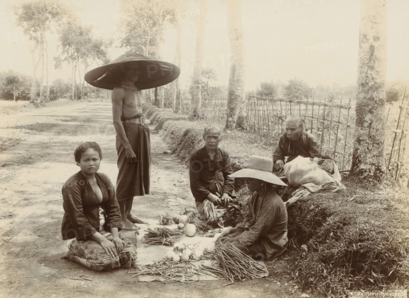 Indonesian women and man with displayed vegetables along the side of the road (former title), Women and man with displayed vegetables along the side of the road, photograph, Part of Travel album with forty photos of the Dutch East Indies., photographer: Onnes Kurkdjian, Dutch East Indies, The, 1912, paper, daylight gelatin silver print, height 177 mm x width 237 mm, selling, A group of people sit and stand on a dirt road beside a grassy embankment and a simple wooden fence with trees lining the roadside; one person stands wearing a large broad hat and a wrapped lower garment, three people sit on the ground around a mat displaying bundles of long slender roots and round vegetables, one seated person wears a hat with a brim and another wears a dark top with a patterned skirt, an older person sits near a large cloth bundle beside the fence, and the scene shows trees, foliage, and a shaded road stretching into the distance.
