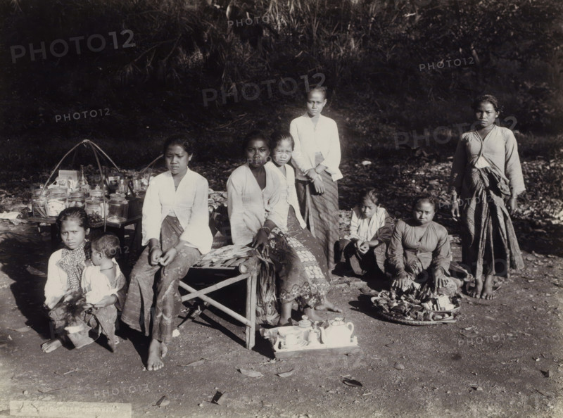 Women and children with goods displayed along the side of the road, Indies women and children with goods displayed along the side of the road (former title), photograph, Part of Travel Album with Forty Photos of the Dutch East Indies., photographer: Onnes Kurkdjian, Dutch East Indies, The, 1912, paper, daylight gelatin silver print, height 176 mm x width 238 mm, merchant, salesman, selling, A group of people arranged outdoors on bare ground in front of a backdrop of dense foliage, with several seated on low stools or the ground and a few standing; most wear patterned lower garments and light-colored tops, and some have scarves or shawls draped around their shoulders; one person holds a small child on their lap; in front of the group are small trays and containers that include jars, a teapot, cups, and other assorted vessels, and a woven tray with wrapped items; two curved supports rise from a low table that holds additional jars and small objects; the lighting creates strong contrasts ...