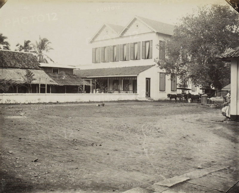 Exterior of the Weltevreden kindergarten, Weltevreden Fröbel school., album page, photograph, Part of Album with photos of Batavia and of the Ursuline sisters., Weltevreden, c. 1890 - c. 1910, photographer, Java, West-Java, Batavia, paper, pasteboard, albumen print, height 243 mm x width 290 mm, nursery school, exterior, representation of a building, A wide dirt foreground with scattered small stones leads to a low white wall in front of a two-story building with a tiled gabled roof and shuttered windows on the upper level, a covered porch with columns and potted plants on the lower level, and a dark doorway visible at ground level; to the left of the building are structures with thatched roofs and palm trees rising behind them, and to the right a large leafy tree partially obscures the building while horse-drawn carriages stand near the tree and figures are visible near the building edge.