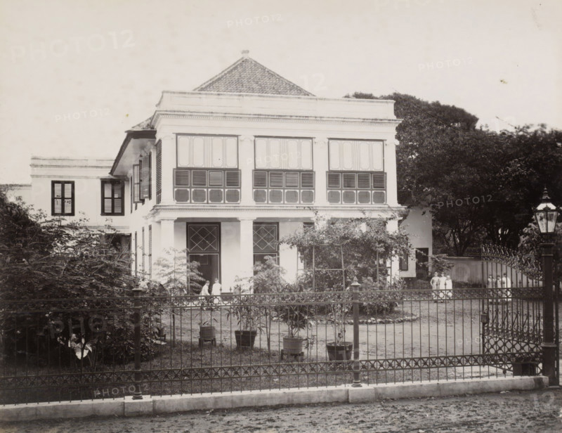 Exterior of the Ursuline Convent of Noordwijk (Batavia), Convent of Noordwijk, front., album page, photograph, Part of Album with photos of Batavia and of the Ursuline sisters., Batavia, c. 1890 - c. 1910, photographer, Java, West-Java, paper, albumen print, height 216 mm x width 278 mm, abbey, monastery, convent, Roman Catholic church, façade (of house or building), A two-story colonial-style house with a tiled, pyramidal roof and a broad upper facade featuring large shuttered windows; the ground level has a covered porch supported by columns and doors with diamond-pane window panels; a wrought-iron fence and gate run across the front of the property with potted plants and small trees placed inside the yard; dense foliage and large trees flank the building on both sides; a tall street lamp stands outside the fence near the gate; several people in light clothing stand near the porch and by the gate.