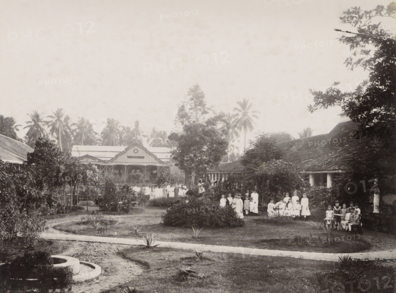 The garden of the convent, Noordwijk., Girls in the convent garden of the Ursuline Sisters of Noordwijk (Batavia), with a building with the inscription 'Bloemendaal' in the background, album page, photograph, Part of Album with photos of Batavia and of the Ursuline Sisters., Batavia, c. 1890 - c. 1910, photographer, Java, West-Java, paper, albumen print, height 216 mm x width 279 mm, garden, abbey, monastery, convent, Roman Catholic church, child, A wide garden scene with a curved dirt path and several small spiky plants in the foreground leading toward a central grassy area with low shrubs and taller bushes, framed by trees and hedges on either side; a group of people, many in light-colored dresses and some seated, are gathered on the right side near a veranda and among the shrubs, with additional figures standing further back along a central walkway; behind them are two large buildings with pitched roofs and decorative gables partially obscured by trees, and tall palm trees rising be...