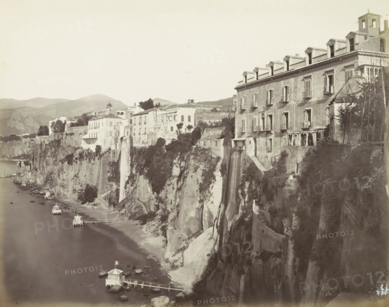 Sorrento!, Houses along a cliff in Sorrento, photograph, Part of Travel album with photos of sights in Naples and the surrounding area., Sorrento, c. 1865 - c. 1875, photographer, paper, albumen print, height 200 mm x width 251 mm, coastal cliff or crag, A seaside cliffside scene showing multiple buildings perched along the cliff edge, with a large multi-story building on the right featuring rows of windows and small balconies and a series of dormer windows on the roof, and a row of smaller light-colored buildings extending leftward along the cliff. Vegetation and trees appear among the buildings and on the cliff face, with steep rocky cliffs dropping to a narrow shoreline. Small pavilions on piers extend into the water below, connected by narrow walkways, and small boats are moored near the shore. Hills and distant mountains form the background under a pale sky.