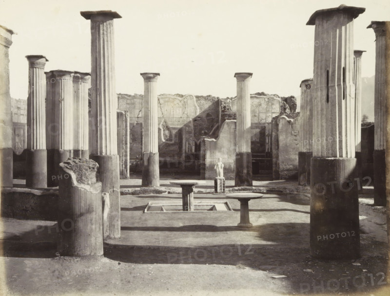 Remains of buildings with a statue in Pompeii, photograph, Part of Travel album with photos of sights in Naples and the surrounding area., Pompeii, c. 1865 - c. 1875, photographer, paper, albumen print, height 195 mm x width 253 mm, column, pillar, architecture, excavation, archaeology, piece of sculpture, reproduction of a piece of sculpture (sculpture), A wide frieze of tall, fluted stone columns rises from a flat paved courtyard, some columns topped with square capitals and some truncated or broken, casting long shadows across the ground; in the foreground three pedestal tables with round tops flank a shallow rectangular basin set into the pavement, and a small statue stands elevated on a stepped plinth near the center background; surrounding walls with rough, weathered surfaces and remnants of plaster enclose the space, with rubble and fractured column stumps visible among the standing columns.