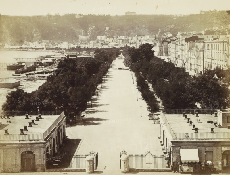 Naples: Royal Garden., View of the Royal Garden in Naples, photograph, Part of a travel album with photos of sights in Naples and the surrounding area., negative number: '4014', c. 1865 - c. 1875, photographer, paper, albumen print, height 198 mm x width 248 mm, public gardens, park (city(-scape) with figures, staffage), A wide elevated view of a broad tree-lined avenue stretching toward a distant promenade, flanked on both sides by rows of dense trees and lined with lamp posts, with a central circular fountain visible along the avenue and a large gated entrance in the foreground; to the left a waterfront area with piers, small boats, and wooden structures extends into the water, while to the right a long row of multi-story classical buildings with uniform façades borders the avenue; rooftops with chimneys and terraces appear in the foreground, and a hillside with scattered buildings and vegetation rises in the background.