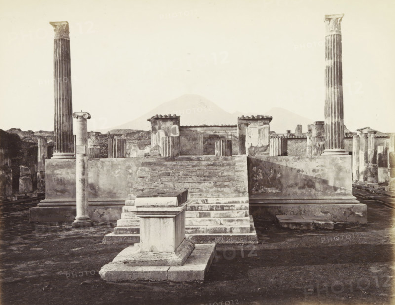 Pompeii: Temple Venus: searches 1817., Remains of the Temple of Venus in Pompeii with Mount Vesuvius in the background, photograph, Part of Travel album with photos of sights in Naples and surroundings., negative number: '6560', c. 1865 - c. 1875, photographer, paper, albumen print, albumendruk, height 208 mm x width 253 mm, column, pillar, architecture, temple, shrine, non-Christian religions, excavation, archeology, A wide view of ancient stone ruins centered on a stepped platform leading up to a raised terrace with low walls and remnants of painted plaster, flanked by tall fluted columns with ornate capitals and shorter broken column stubs, with a solitary slender column standing in the foreground on a square base and a rectangular plinth positioned at the base of the steps, all set on a dark textured ground and with distant low hills visible against a pale sky.