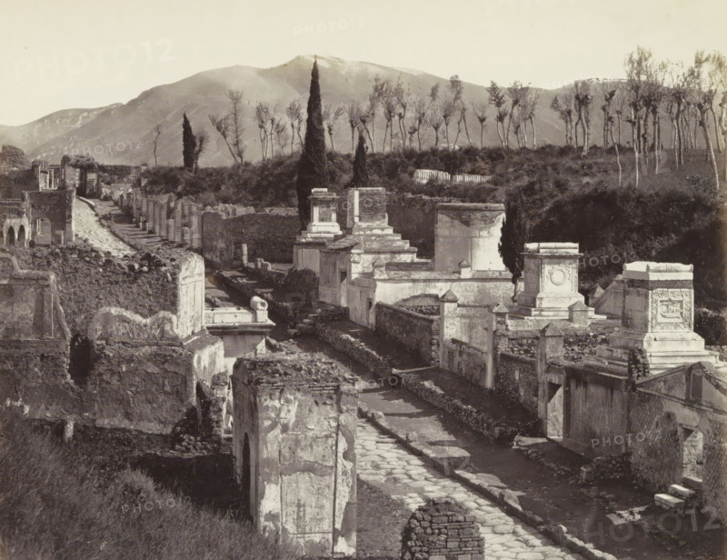 Pompei: Rue des tombeau fouilles 1763, Remains of buildings on the Street with Tombs (Via delle Tombe) in Pompei, with Mount Vesuvius in the background, photograph, Part of Travel album with photos of sights in Naples and the surrounding area., negative number: '4207', Pompeii, c. 1865 - c. 1875, photographer, paper, albumen print, albumendruk, height 201 mm x width 248 mm, street, grave, tomb, excavation, archaeology, A wide grayscale image of an ancient street lined with stone and plaster ruins, showing broken walls, column bases, and rectangular tomb-like structures with decorative reliefs. A paved roadway runs through the center foreground toward the background, flanked by low retaining walls and narrow passageways between structures. Tall slender trees and cypress-like evergreens stand on a raised embankment behind the ruins, with a line of taller bare-branched trees further back. Rolling hills and a mountain rise in the distant background beneath a pale sky. The scene shows worn ...