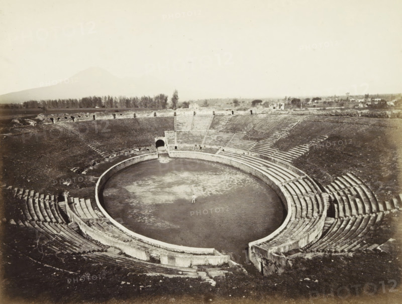 Interior of the amphitheater of Pompeii, Pompei: Amphitheatre. (pour 20 mille spectators) excavations 1813., photograph, Part of Travel Album with photos of sights in Naples and the surrounding area., negative number: '4209', Pompeii, c. 1865 - c. 1875, photographer, paper, albumen print, height 203 mm x width 252 mm, theater (building) - AA - open-air performances, A broad view of a circular amphitheater with concentric stone seating rising from a central flat arena, showing worn steps and curved retaining walls; a single small figure stands in the center of the arena; the seating tiers show gaps, diagonal stairways, and entry passages, one of which is a dark arched tunnel opening onto the arena; beyond the amphitheater, a low stone perimeter and a line of trees stretch across the horizon, with scattered buildings and distant vegetation under a pale sky.