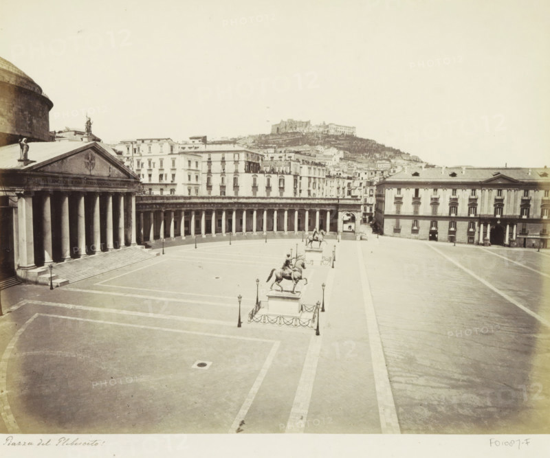Piazza del Plebescito with equestrian statues in Naples, Piazza del Plebescito, photograph, Part of Travel album with photos of sights in Naples and surroundings., Naples, c. 1865 - c. 1875, photographer, paper, albumen print, albumendruk, height 199 mm x width 252 mm, equestrian statue, square, circus, etc. (city(-scape) with figures, staffage), A wide pale plaza with tiled paving is flanked by a large classical building with a columned portico and a triangular pediment on the left and a long colonnade curving across the middle ground, with multi-story buildings rising behind them; three equestrian statues mounted on raised plinths stand along the plaza center, each surrounded by short posts and chains and paired with tall street lamps; a broad open roadway runs along the right side toward a rectangular building with arched entrances and evenly spaced windows; in the distant background a hillside with layered buildings and a prominent fortress-like structure sits beneath a pale sky.