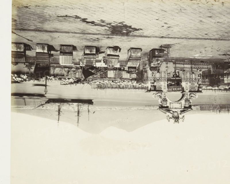 Harbor of Naples in the Santa Lucia neighborhood with oyster vendors' stalls in the foreground, S. Lucia. La Plage d'huîtres, photograph, Part of Travel Album with photos of sights in Naples and the surrounding area., photographer: attributed to Giorgio Sommer, Naples, c. 1865 - c. 1875, Attributed to, paper, albumen print, height 199 mm x width 250 mm, stall, booth (, market), harbor (view, vehicle, ship, etc.), An upside down black and white photograph showing a waterfront scene with a paved promenade, a row of wooden market stalls and carts, a wrought iron fence with decorative stone pillars and sculptural details, scattered crates and awnings, a lamppost, piles of objects near the water’s edge, reflections of masts and rigging in the water, and distant hills on the horizon.