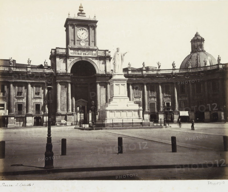 Piazza Dante in Naples with statue of Dante and the Lyceum Vittorio Emanuele, Piazza S. Spirito!, photograph, Part of Travel album with photos of sights in Naples and surroundings., Naples, c. 1865 - c. 1875, photographer, paper, albumen print, height 194 mm x width 253 mm, monument, statue, square, circus, etc. (city(-scape) with figures, staffage), secondary education (building, education, tuition), A wide plaza with a paved foreground leads to a curved colonnaded building topped by a balustrade with multiple standing statues, a central monumental arch flanked by tall columns, and a square clock tower rising above the arch; in front of the arch stands a large tiered pedestal supporting a draped standing statue with one arm extended, and to the right a patterned domed roof with a lantern rises behind the balustrade; a tall ornate lamppost is visible in the foreground, short bollards dot the plaza, and a few small figures stand near the building and on the open square.