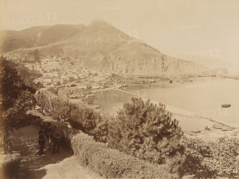 View of the port of Oran, Oran. Le Djabel Mourdjadjo et le Port, vue prize de la promenade de Letang, album page, photograph, This photo is part of an album., number: '204', photographer: Étienne Neurdein, c. 1880 - c. 1890, paper, albumen print, albumendruk, height 207 mm x width 272 mm, harbour, city-view in general, 'veduta', mountains, North Africa, Oran, A sepia-toned panoramic view showing a coastal town at the foot of a large rocky hill, with clustered buildings and docks along a calm bay, a breakwater extending into the water, small boats moored near the shore and one floating further out, steep cliffs rising behind the harbor, terraced slopes and scattered structures on the hillside, foreground vegetation including trees and hedges, and three men dressed in dark coats and hats standing on a shaded path overlooking the scene.