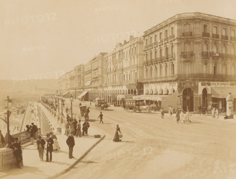 View of Boulevard de la Republique in Algiers, Alger. - Boulevard de la Republique, album page, photograph, This photo is part of an album., number: '284', photographer: Étienne Neurdein, Algiers, 1880 - 1890, North Africa, paper, albumen print, albumendruk, height 208 mm x width 273 mm, historical persons, avenue, boulevard, promenade, esplanade, A wide street scene with a long row of ornate multi-story buildings featuring balconies and arched ground-level openings along the right side, horse-drawn carriages and a larger horse-drawn omnibus on the roadway, groups of people walking and standing on the sidewalk and promenade, lampposts lining the elevated promenade with a balustrade, awnings and canopies over some shopfronts, and distant low hills visible in the background.