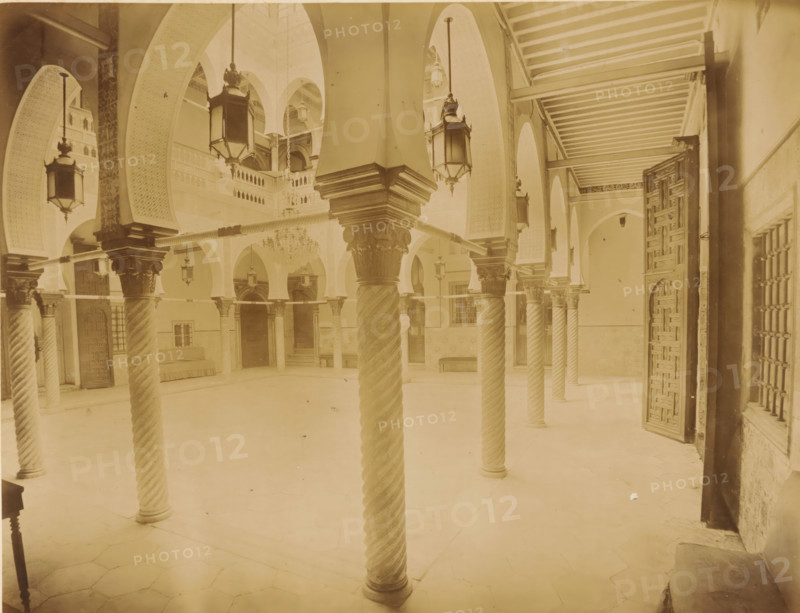 Courtyard of Palais des Raïs in Algiers, Alger. Interior du Palais du Gouverneur, album page, photograph, This photo is part of an album., number: '224', photographer: Étienne Neurdein, c. 1880 - c. 1900, paper, albumen print, albumendruk, height 210 mm x width 272 mm, arch, archivolt, architecture, column, pillar, palace, North Africa, Algeria, A vaulted interior space with tall arches and ornate carved columns featuring twisted shafts and decorated capitals, hanging lanterns suspended from the arches, an open carved wooden door on the right, rows of wooden beams across the ceiling at the right, a chandelier suspended in the central background, patterned wall panels and small balconies above, tiled floor, benches and doorways along the far walls, and decorative latticework on the right window.