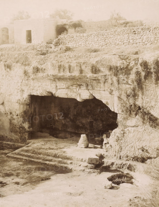 Courtyard of the Tombs of the Kings in Jerusalem, Tombeau des Rois, cour intérieure -Jérusalem, photograph, Part of Travel Album with photos of Jerusalem and other biblical places., photographer: Maison Bonfils, Jerusalem, c. 1867 - c. 1895, pasteboard, paper, albumen print, height 223 mm x width 282 mm, grave, tomb, A weathered stone cliff face with a dark cave opening sits beneath a ledge carved with decorative rectangular and circular motifs, above which a rough terrace of grass and low vegetation gives way to a dry stone wall; to the left on the terrace a small square stone structure with an open doorway and nearby arched recesses is visible, and scattered stones and a short stepped platform lead down from the cave entrance to a flat, worn ground where a small stacked stone formation and several loose blocks rest, all captured in a soft, high-contrast monochrome tone.