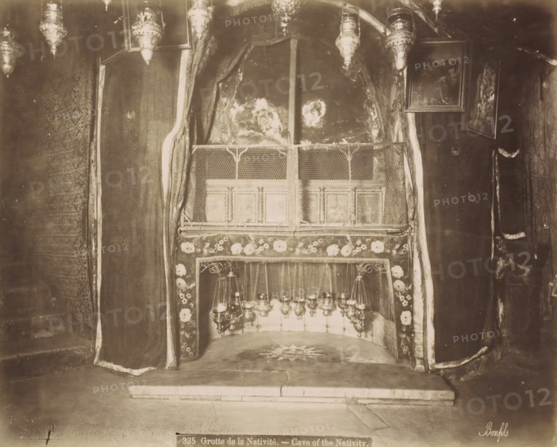Grotte de la Nativite.- Cave of the Nativity., Nativity Cave in the Church of the Nativity in Bethlehem, photograph, Part of Travel Album with Photos of Jerusalem and Other Biblical Places., negative number: '335', photographer: Maison Bonfils, Bethlehem (Israel), c. 1867 - c. 1895, Palestine, pasteboard, paper, albumen print, height 225 mm x width 283 mm, interior of church, cave, grotto, A sepia-toned interior scene showing a small recessed alcove framed by heavy draped curtains with patterned borders and floral trim, a decorative metal grille across the upper front of the recess, hanging ornate lamps suspended inside the lower portion, a stone floor with a star-like design in front of the alcove, textured walls and framed pictures mounted on the wall to the right, and additional ornate hanging lamps visible along the top edge.