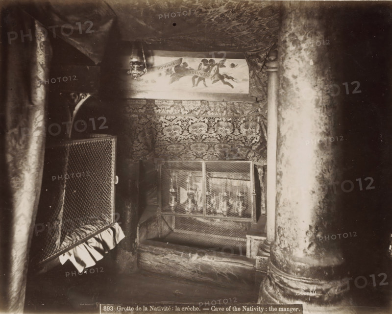 Crib in the Cave of the Nativity in Bethlehem, Grotte de la Nativite: la crèche.- Cave of the Nativity: the manger., photograph, Part of Travel Album with Photos of Jerusalem and Other Biblical Places., negative number: '893', photographer: Maison Bonfils, Bethlehem (Israel), c. 1867 - c. 1895, Palestine, pasteboard, paper, albumen print, height 222 mm x width 282 mm, cradle, crib, A dim interior scene with patterned fabric draped across the ceiling and walls, a hanging ornate lamp near the top center, a painted panel showing figures and clouds partially visible above the fabric, a decorative tapestry with intricate motifs covering the back wall, a metal mesh enclosure with objects behind it in the middle foreground, heavy textured columns on the right and left edges, and a ruffled cloth hanging from a mesh panel on the left.