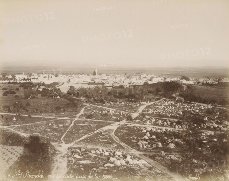 View of Ramla in Israel, Ramallah and surrounding land, seen from a tower (former title), Rameleh, vue générale prise de la tour, photograph, View of Ramla with the Plain of Sharon and the Mountains of Judea, in the foreground an Islamic cemetery. Part of Travel Album with photographs of Jerusalem and other biblical places., negative number: '241bis', photographer: Maison Bonfils, Ramallah, c. 1867 - c. 1895, Palestine, pasteboard, paper, albumen print, height 225 mm x width 283 mm, prospect of city, town panorama, silhouette of city, Ramla, A sepia-toned aerial view shows a broad landscape with a foreground of rocky, uneven ground marked by winding dirt paths and scattered stone markers forming small rectangular shapes, some grouped and some isolated; patches of low vegetation and occasional trees dot the area. In the middle distance a belt of darker trees and denser foliage separates the foreground from a low-lying town of light-colored buildings with flat and pitched roofs, among wh...