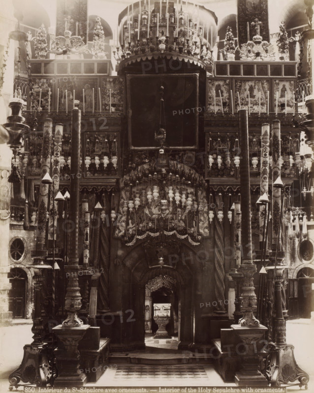 Intérieur du St.-Sépulcre avec ornements. - Interior of the Holy Sepulcher with ornaments., Interior of the Church of the Holy Sepulcher in Jerusalem, photograph, Part of Travel album with photos of Jerusalem and other biblical places., inscription: '...ves No. 74', negative number: '850', photographer: Maison Bonfils, Jerusalem, c. 1867 - c. 1895, pasteboard, paper, albumen print, height 282 mm x width 225 mm, interior of church, Church of the Holy Sepulchre, A large ornate interior view filled with carved columns, tall decorative poles, hanging lamps, and intricately patterned panels; a central dark archway framed by twisted pillars leads inward toward a smaller altar piece visible through the opening, while above the arch are rows of ornate metalwork, suspended lamps, and sculpted reliefs; flanking the central opening are tall painted columns and carved pedestals topped with finials and cone-shaped covers, and the upper portion shows framed panels with figural images and floral deco...