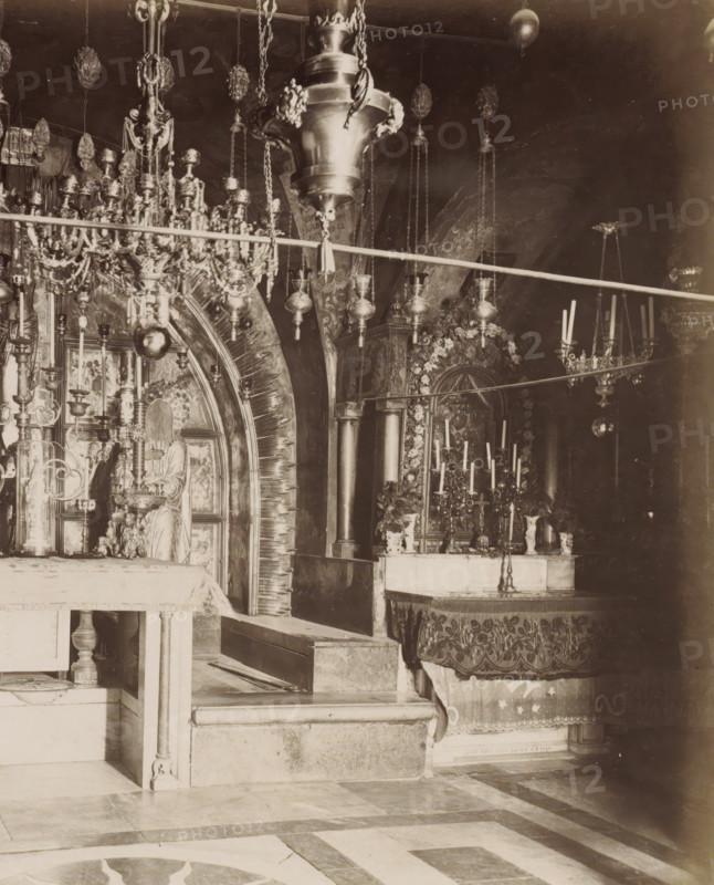 XII et XIII Stations, le Calvaire, autel grec.- places., negative number: '270', photographer: attributed to Maison Bonfils, Church of the Holy Sepulchre, c. 1867 - c. 1895, Jerusalem, Attributed to, pasteboard, paper, albumen print, height 224 mm x width 283 mm, altar, stations of the cross, Church of the Holy Sepulcher, A sepia-toned interior photograph showing an ornate religious space with hanging lamps and elaborate metal chandeliers suspended from the ceiling, a decorative archway with radiating carved elements, carved columns, candleholders with tall candles, an altar draped with patterned cloth, a statue or carved figure seated within a niche, floral garlands around a framed decorative panel, intricate metalwork and chains, stone steps and a tiled floor with geometric patterns.