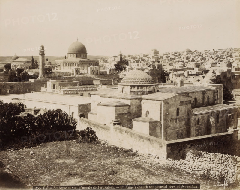 Eglise Ste-Anne et vue générale de Jérusalem.- St Anne's church and general view of Jerusalem., View of Jerusalem with St. Anne's Church in the foreground, photograph, Part of Travel album with photos of Jerusalem and other biblical places., negative number: '255', photographer: Maison Bonfils, Jerusalem, c. 1867 - c. 1895, pasteboard, paper, albumen print, height 226 mm x width 283 mm, prospect of city, town panorama, silhouette of city, church (exterior), St. Anne's Church (Jerusalem), A wide sepia-toned cityscape with a large domed building prominently near the center, several smaller domes and towers scattered across the view, a tall cylindrical minaret with a balcony rising in front of the domed complex, stone buildings with flat roofs and arched windows filling the foreground and stretching into the distance, low walls and terraces creating layered levels, patches of trees and vegetation interspersed among the structures, a rough rocky foreground leading up to the built area, and...
