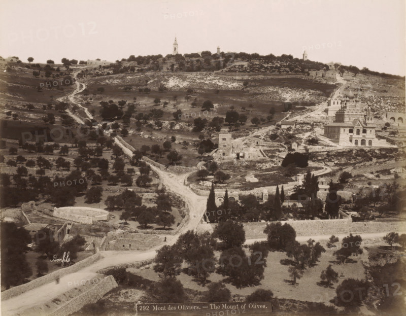 View of the Mount of Olives in Jerusalem, Mont des Oliviers. - The Mount of Olives., photograph, Part of Travel Album with photos of Jerusalem and other biblical places., negative number: '292', photographer: Maison Bonfils, Jerusalem, c. 1867 - c. 1895, pasteboard, paper, albumen print, height 223 mm x width 281 mm, mountains (landscape with figures, staffage), Mount of Olives, A wide sepia-toned landscape showing a hilly terrain with scattered trees and winding dirt paths leading upward, stone terraces and low walls breaking the slopes, a cluster of buildings on the right including a multi-domed ornate structure and adjacent rectangular buildings, ruins and small stone structures dotting the midground, taller tower-like structures visible on the ridge in the distance, and a foreground with more trees, stone enclosures, and a curved road.