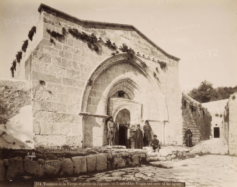 Tomb of the Virgin Mary and the Cave of the Agony in Jerusalem, Tombeau de la Vierge et grotte de l'agonie. - Tomb of the Virgin and cave of the agony., photograph, Part of Travel album with photos of Jerusalem and other biblical places., negative number: '314', photographer: Maison Bonfils, Jerusalem, c. 1867 - c. 1895, Maison Bonfils, photographer, pasteboard, paper, albumen print, height 225 mm x width 284 mm, church (exterior), grave, tomb, cave, grotto, A stone building facade with a large arched entrance and smaller arched recesses, weathered block masonry and patches of vegetation growing on and along the upper walls; a cobbled foreground with low stone steps and a raised stone platform; several men in long garments and hats gathered near the entrance, one seated on the steps and others standing; a narrow passage to the right lined by a high stone wall topped with foliage and a distant dark doorway; pale sky above.