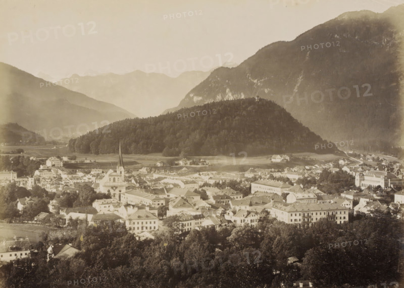 Ischl vom Gassner, View of Ischl and surrounding mountains from the Gassner, photograph, Part of travel album with photos of sights in Germany and Austria and of works of art., negative number: '345', Ischl, c. 1870 - c. 1900, photographer, Upper Austria, paper, albumen print, height 198 mm x width 267 mm, prospect of city, town panorama, silhouette of city, A wide sepia-toned view of a town nestled at the base of a densely wooded hill and surrounded by larger mountains, showing a foreground of clustered buildings with pitched roofs and varied architectural styles, interspersed with trees and open spaces, and a prominent church with a tall pointed spire rising above nearby structures, while the midground features a tree-covered hill topped by a small tower, and the background reveals layered mountain slopes fading into the sky.