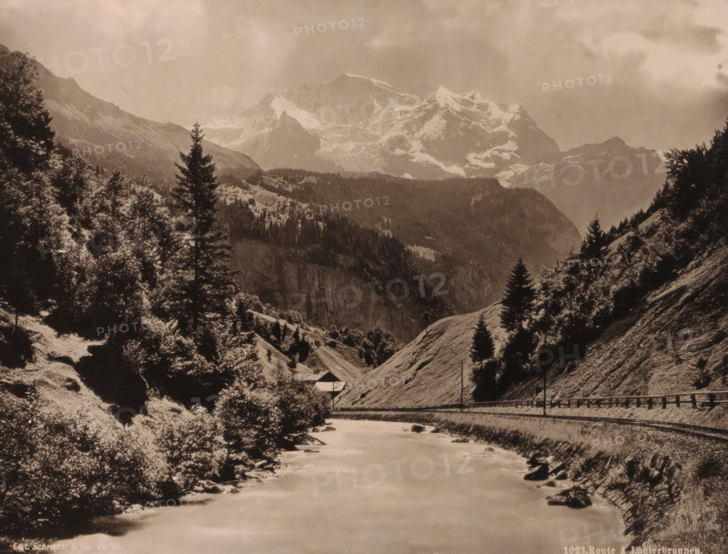 Route to Lauterbrunnen., Mountain landscape with the railway from Interlaken to Lauterbrunnen, album page, photograph, Part of Travel album with sights in Germany, Austria, Switzerland, Luxembourg and Belgium., number: '1023', photographer: Schroeder & Cie., c. 1880 - in or before 1899, ink, materials by function, paper, printing ink, collotype, photogravure, height 210 mm x width 275 mm, railway, train, mountain-range, snow peaks, A sepia-toned mountain scene with a river flowing toward the foreground, flanked by grassy slopes and rocky banks. Evergreen trees and clusters of leafy trees rise on both sides of the valley, with taller conifer silhouettes standing prominently against the sky. A narrow road or rail line runs along the right bank, bordered by a low fence and a few utility poles. A small building with a sloped roof sits near the base of a hillside in the midground. Layered ridges and forested hills lead back to a jagged, snow-covered mountain range under a cloudy sky.