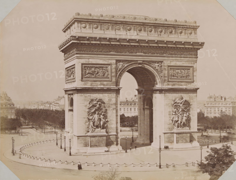 View of the Arc de Triomphe, album page, photograph, This photo is part of an album., retailer's mark: 'E. Hautecoeur, Estampes Photographies, 35 Avenue de l'Opéra, 35, Paris', inscription: '456B Arc de Triomphe. Ed. H.edit. Paris', photographer: Edouard Hautecoer (possibly), Paris, 1890 - 1898, paper, albumen print, height 258 mm x width 355 mm, triumphal arch, Arc de Triomphe, A large stone triumphal arch dominates the image, showing a central arched passage and flanking massive piers with high relief sculptures attached to their faces; decorative friezes and carved circular medallions run along the upper entablature, and rectangular relief panels sit above the arch on each pier. The arch opening reveals a coffered underside and a distant view through to buildings and trees beyond. A paved plaza surrounds the monument, edged by a low balustrade with chained posts and street lamps, and a few small groups of people and a solitary figure are visible near the base of the arch. Rows of tr...
