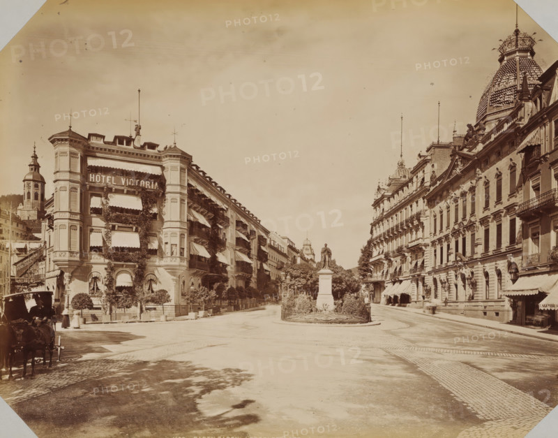 Leopoldplatz in Baden-Baden, album page, photograph, This photo is part of an album., inscription: '1673 Baden-Baden Leopoldplatz LL', photographer: LL, Baden-Baden, 1890 - 1899, Baden-Württemberg, paper, albumen print, albumendruk, height 258 mm x width 355 mm, square, circus, etc. (city(-scape) with figures, staffage), A wide city street flanked by multi-story ornate buildings with decorative facades and awnings stretches toward a central statue on a pedestal surrounded by low hedges and trees, the left building covered in climbing plants and window awnings and the right building featuring balconies and a large domed roof with patterned tiles, lamp posts and flagpoles rising above the rooftops, a cobblestone roadway and sidewalk in the foreground, a horse-drawn carriage and a few pedestrians visible near the left edge, and distant towers and architectural details visible down the street beneath a pale sky.