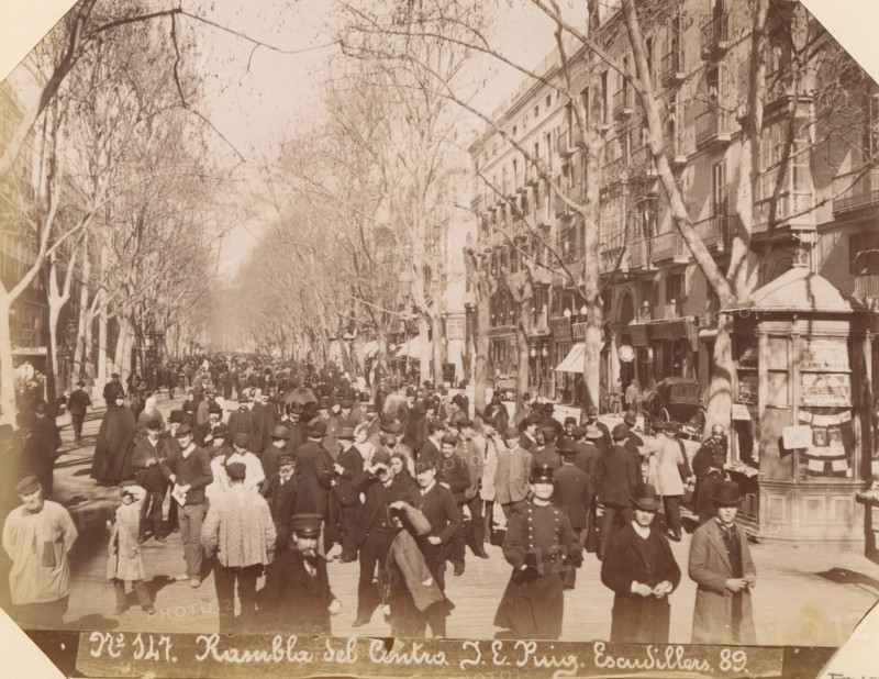 Rambla del Centro, Street view at Rambla dels Caputxins in Barcelona, album page, photograph, This photo is part of an album., annotation: 'Barcelona - Rambla de las Flores y del Centro', number: '147', photographer: JE Puig, Barcelona (city), c. 1885 - in or before 1892-04-09, Catalonia, paper, albumen print, height 163 mm x width 210 mm, street, A wide tree-lined street filled with many pedestrians wearing period clothing, including coats, hats, and long dresses, stretches into the distance; tall bare-branched trees line both sides of the street and cast shadows across the scene, while multi-story buildings with balconies and shopfronts run along the right side; a small kiosk or rounded structure with posters attached stands near the right foreground, and numerous streetlamps and awnings are visible among the storefronts; the crowd includes men, women, and children walking or standing, some grouped together and some facing toward the camera, with a few horse-drawn vehicles partially ...