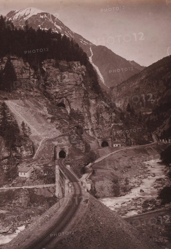 Gotthard Railway Faido, View of the Gotthard Railway at Faïdo, album page, photograph, This photo is part of an album., annotation: 'Gotthard railway at Faïdo', number: '12894', photographer: Giorgio Sommer, c. 1885 - c. 1892, paper, albumen print, height 199 mm x width 254 mm, railway, train, mountains, Faido, A sepia-toned landscape showing a rocky mountain slope with patches of snow near the peak, dense dark tree cover on parts of the slopes, and exposed rock faces. A river with white foamy water runs through a rocky riverbed alongside a narrow road or path that winds up the slope. Stone tunnels with dark arch openings are built into the rock next to a curved section of road or rail, and small stone structures or buildings sit near the road and tunnel entrances. The sky appears clear and pale above the mountains, and the image is framed by a light border along the top and side.