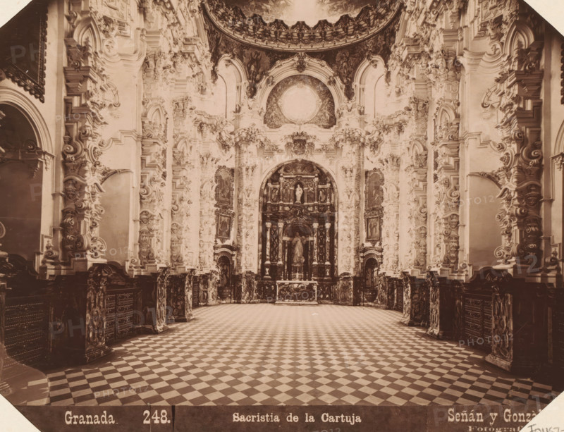 Interior of the sacristy of the Real Monasterio de La Cartuja in Granada, Granada. Sacristia de la Cartuja, album page, photograph, This photo is part of an album., annotation: 'Granada - Sacristie der Cartuga', number: '248.', photographer: Señan y Gonzalez, c. 1885 - in or before 1892-03-12, paper, albumen print, height 159 mm x width 208 mm, parts of church interior: sacristy, Andalusia, Granada, A large ornate interior space with a wide checkered floor leading toward a richly decorated altar niche, the walls and columns filled with elaborate carved and sculpted ornamentation including scrolls, floral motifs, and framed panels, high arched openings and recessed alcoves, a central arched area containing statues and layered architectural elements, decorative molding and relief work rising to a domed or curved ceiling with painted or darkened panels, dark base panels and carved cabinetry lining the lower walls, and soft sepia-toned lighting across the whole scene.
