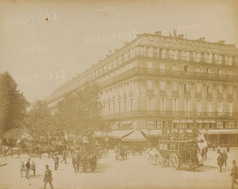 View of the Boulevard des Capucines with horse-drawn trams and the Grand Hotel in Paris, photograph, Part of a travel album with photographs of sights in Sweden, Norway, Denmark, Bavaria, Milan, and Paris., Paris, c. 1870 - c. 1890, photographer, paper, albumen print, height 220 mm x width 278 mm, street, hotel, hostelry, inn, four-wheeled, animal-drawn vehicle, eg: cab, carriage, coach, A wide street scene showing a large multi-story corner building with rows of tall windows and shuttered upper windows, ornate balconies, and a sloped mansard roof with chimneys and rooftop railings, trees lining the street in front of the building, horse-drawn carriages and coaches moving along the roadway, a double-decker horse bus with an open upper deck, people walking and standing on the pavement and near the vehicles, awnings extending from the ground floor of the building, decorative architectural details on the building facade, and a hazy sky above.