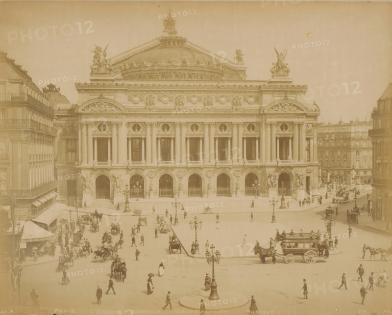 View of the Opéra Garnier in Paris, PARIS L'Opéra, Académie Nationale de Musique, photograph, Part of Travel album with recordings of sights in Sweden, Norway, Denmark, Bavaria, Milan and Paris., number: '445.A', photographer: H, Paris, c. 1870 - c. 1890, H, photographer, paper, albumen print, albumendruk, height 219 mm x width 275 mm, opera house, Opéra de Paris, A wide sepia photograph of a classical stone building with a large central dome and ornate sculptures along the roofline, flanked by symmetrical facades with tall columns and arched openings; the facade features decorative reliefs and circular medallions above the windows, and statues stand in niches near the entrance arches. In front of the building is a broad open plaza with multiple ornate street lamps and scattered groups of pedestrians walking or standing, some carrying umbrellas, and several horse-drawn carriages and carts moving across the square. Buildings with uniform facades and balconies line the sides of the plaza...