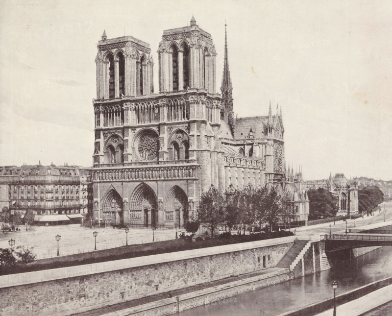 Notre-Dame of Paris, album page, photomechanical print, This print is part of an album., maker: Angerer & Göschl, C., 1880 - 1900, paper, autotype, height 201 mm x width 248 mm, river, church (exterior), Paris, Notre-Dame (Paris), A large Gothic cathedral façade dominates the image with two tall square towers, a central circular rose window, ornate arched doorways, and rows of vertical columns and sculptural details. A slender pointed spire rises behind the towers above the roofline. Trees and a low balustrade sit in front of part of the building, and a riverside embankment with stone steps, a retaining wall, and a bridge span the foreground. To the left and behind the cathedral are multi-story buildings with regular windows and mansard roofs, and lamp posts line the open paved area between the cathedral and the embankment. The scene is shown in a monochrome, grainy tone.