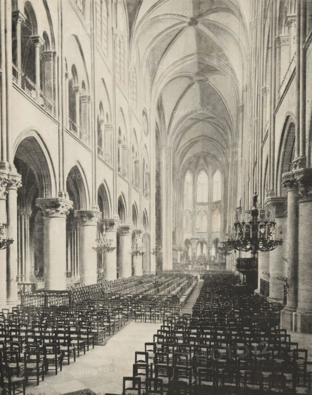 Interior of Notre-Dame in Paris, album page, photomechanical print, This print is part of an album., maker: Angerer & Göschl, C., 1880 - 1900, paper, autotype, height 250 mm x width 198 mm, interior of church, Paris, Notre-Dame (Paris), A large interior space with tall vaulted ceilings and a long central aisle lined with rows of wooden chairs, flanked on one side by a series of massive stone columns supporting rounded arches and an upper gallery with smaller arches and windows, and on the other side by similar columns and architectural details; ornate chandeliers hang from the ceiling and are positioned along the side aisles, decorative capitals sit atop the columns, and light filters through tall arched windows at the far end, illuminating the distant choir area and altar rail.
